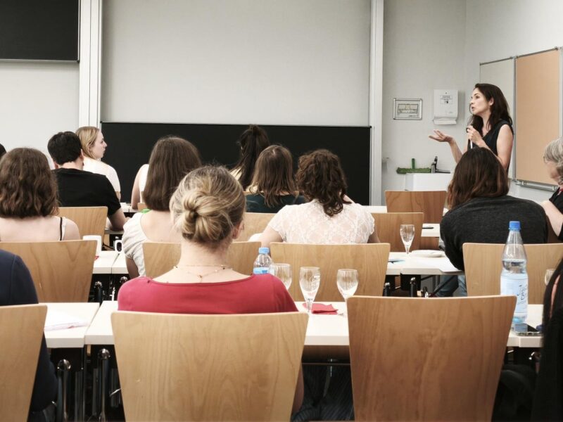 A group of pre-service teachers photographed from behind, sitting in a classroom, listening to the person speaking at the front.