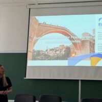 A person holding a presentation in front of a projection screen showing the bridge of Mostar, EUPeace's logo.