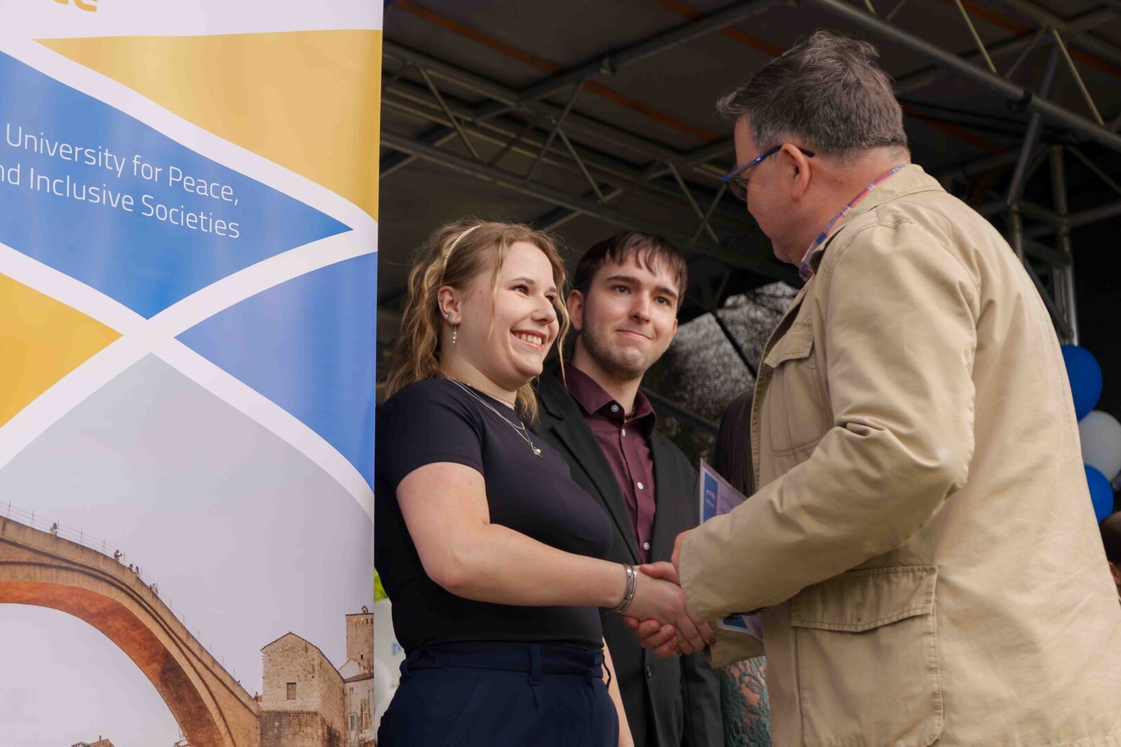 A man shaking a student's hand while congratulating her and presenting a certificate. Another student next to her looks toward the man as the certificate is being given.