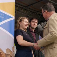 A man shaking a student's hand while congratulating her and presenting a certificate. Another student next to her looks toward the man as the certificate is being given.
