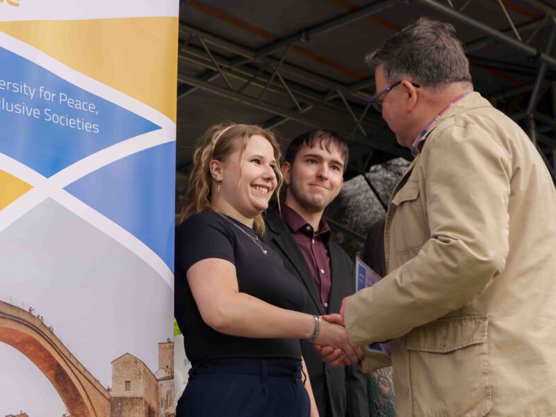 A man shaking a student's hand while congratulating her and presenting a certificate. Another student next to her looks toward the man as the certificate is being given.