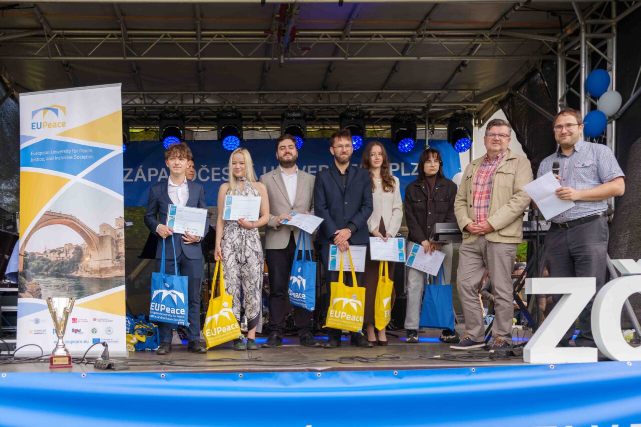 A group of students and staff standing on a stage next to an EUPeace banner, holding their European Track Certificates and EUPeace tote bag.