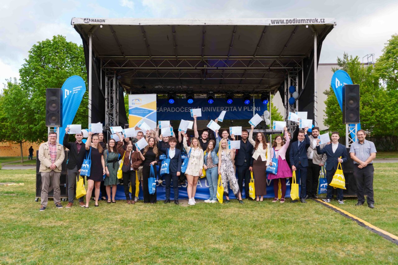 A group of students and staff in front of a stage posing for a group photo, holding their EUTrack Certificates in the air.