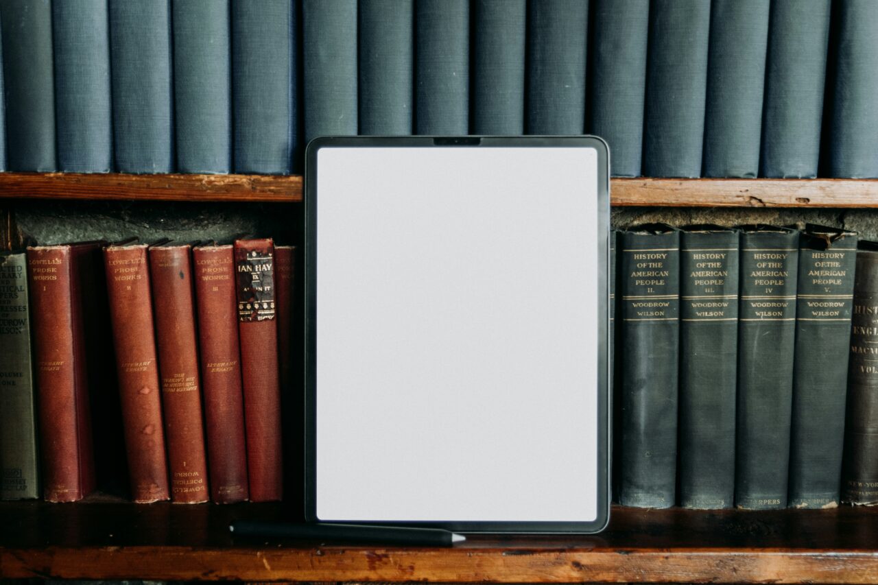 A tablet with a blank white screen stands upright on a wooden bookshelf, surrounded by rows of old hardcover books including volumes of Lowell's Prose Works and Woodrow Wilson's History of the American People.