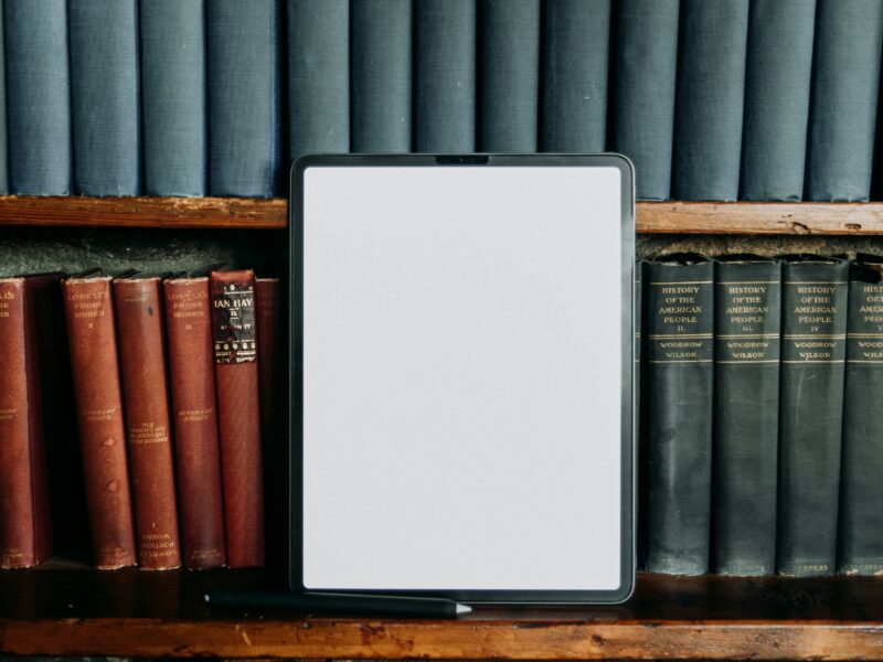 A tablet with a blank white screen stands upright on a wooden bookshelf, surrounded by rows of old hardcover books including volumes of Lowell's Prose Works and Woodrow Wilson's History of the American People.