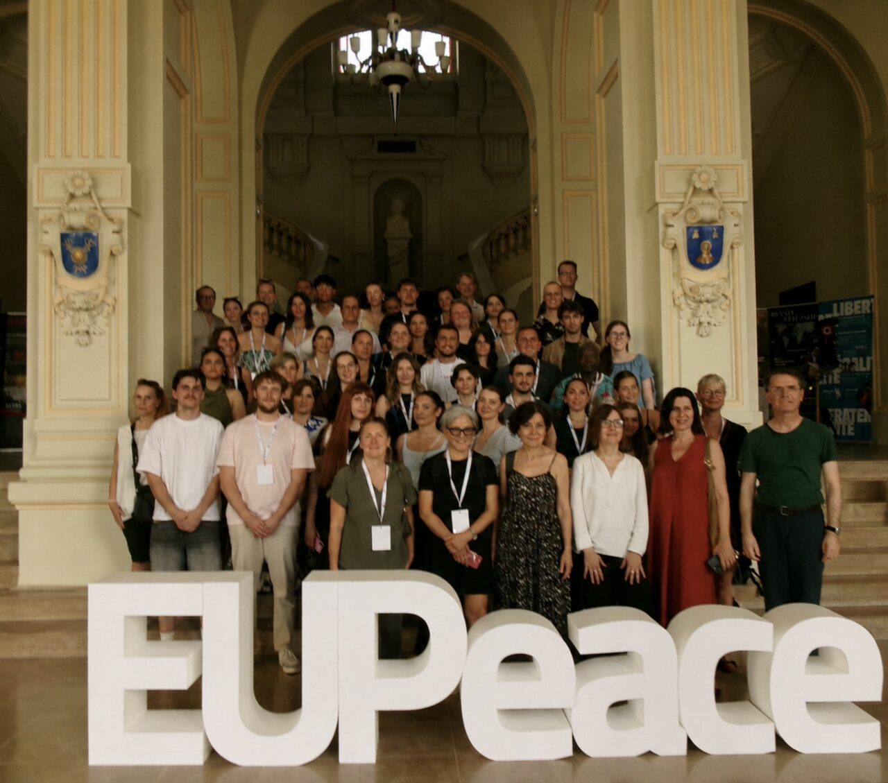 A large group of participants poses for a group photograph in front of large white three-dimensional EUPeace lettering, inside an ornate building with arched columns and a grand staircase.
