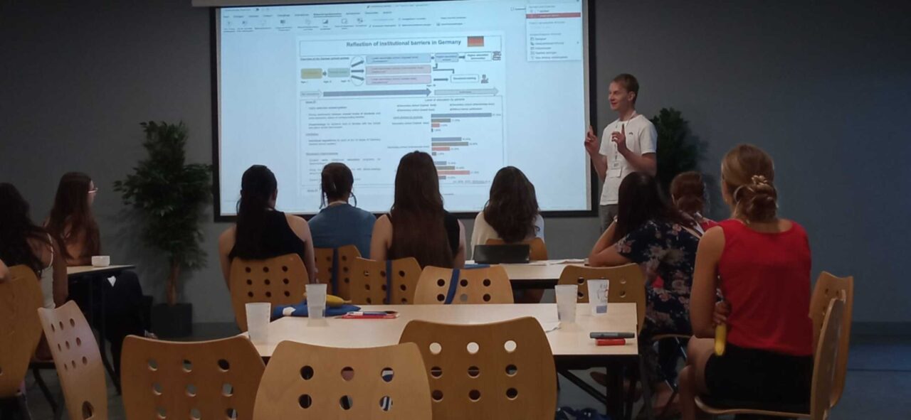 A presenter stands beside a large projection screen displaying a slide titled "Reflection of institutional barriers in Germany", addressing a group of seated participants during the EUPeace Summer School 2026 at the University of Limoges.