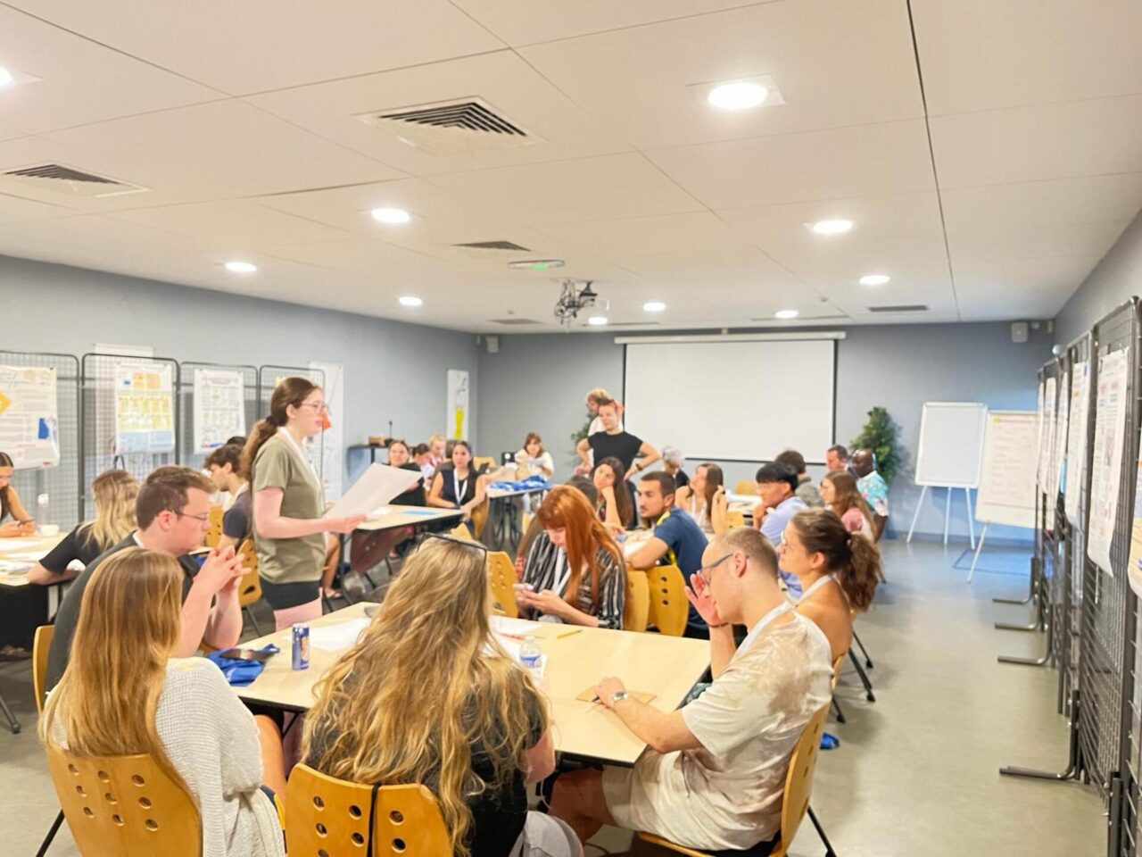 A large group of participants seated at tables during a collaborative workshop session at the EUPeace Summer School 2026. A facilitator stands and addresses the group, with poster displays and a flipchart visible along the walls.