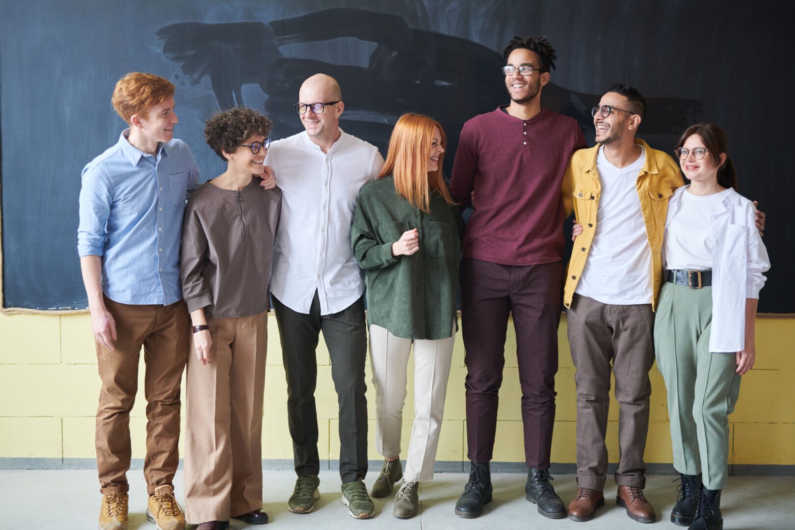 Seven smiling colleagues stand arm in arm in a row in front of a chalkboard wall, dressed in casual business attire.