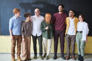 Seven smiling colleagues stand arm in arm in a row in front of a chalkboard wall, dressed in casual business attire.