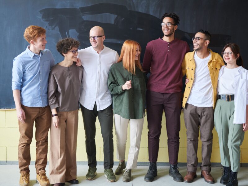 Seven smiling colleagues stand arm in arm in a row in front of a chalkboard wall, dressed in casual business attire.