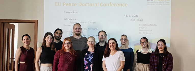 A group of eleven conference participants pose for a photo in front of a projected presentation slide reading "EUPeace Doctoral Conference."