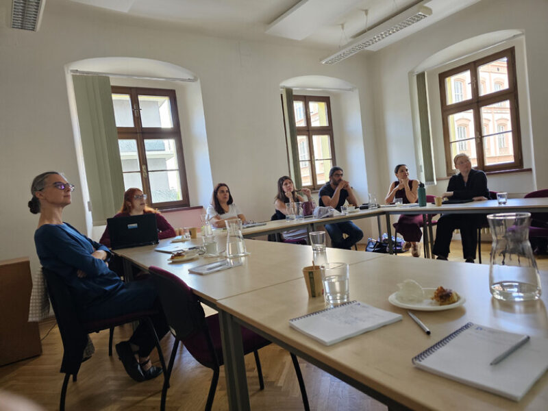 A group of seven conference participants sit attentively at tables in a bright seminar room with arched windows, listening to a presentation.