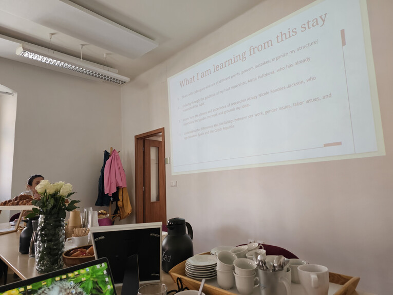 A seminar room with a projected slide titled "What I am learning from this stay," with refreshments and a laptop visible in the foreground.