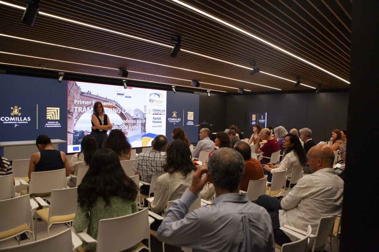 An audience sitting on chairs, photographed from behind and listening to someone speaking in the front row. A woman is standing at the front with a big screen behind her.