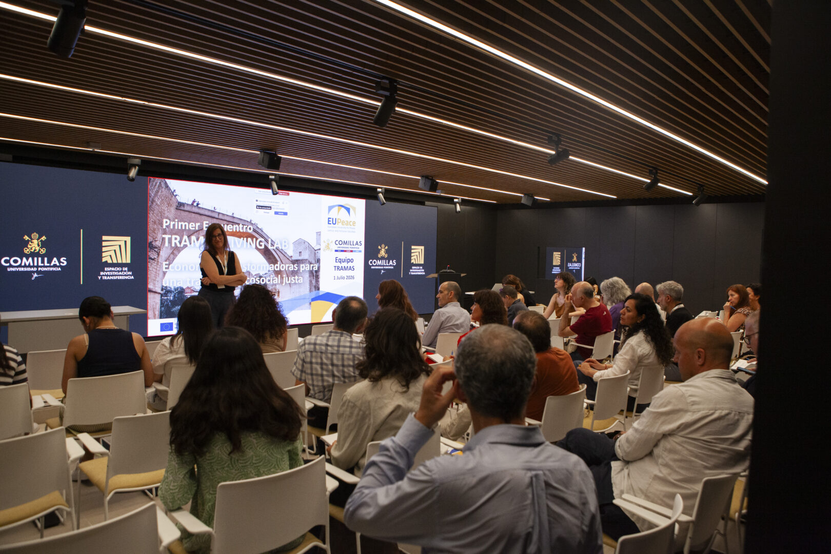 An audience sitting on chairs, photographed from behind and listening to someone speaking in the front row. A woman is standing at the front with a big screen behind her.