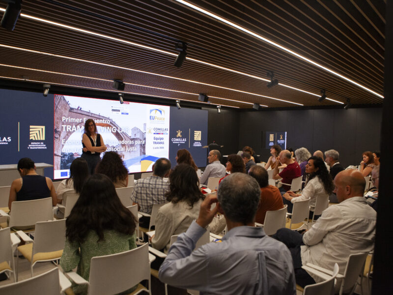 An audience sitting on chairs, photographed from behind and listening to someone speaking in the front row. A woman is standing at the front with a big screen behind her.