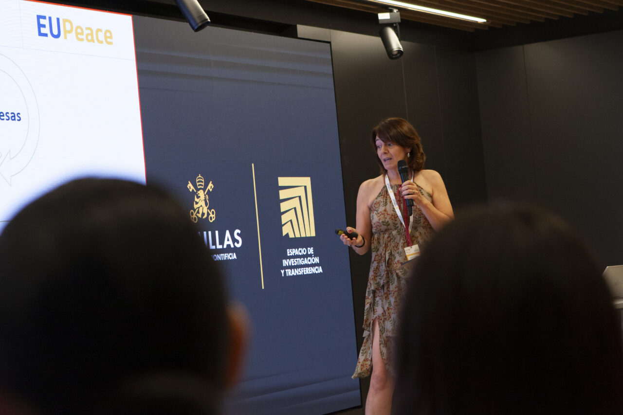 A woman standing in front of a big screen holding a wireless presenter and speaking into a microphone in her other hand. The blurry backs of two heads are positioned in the two bottom corners of the image,