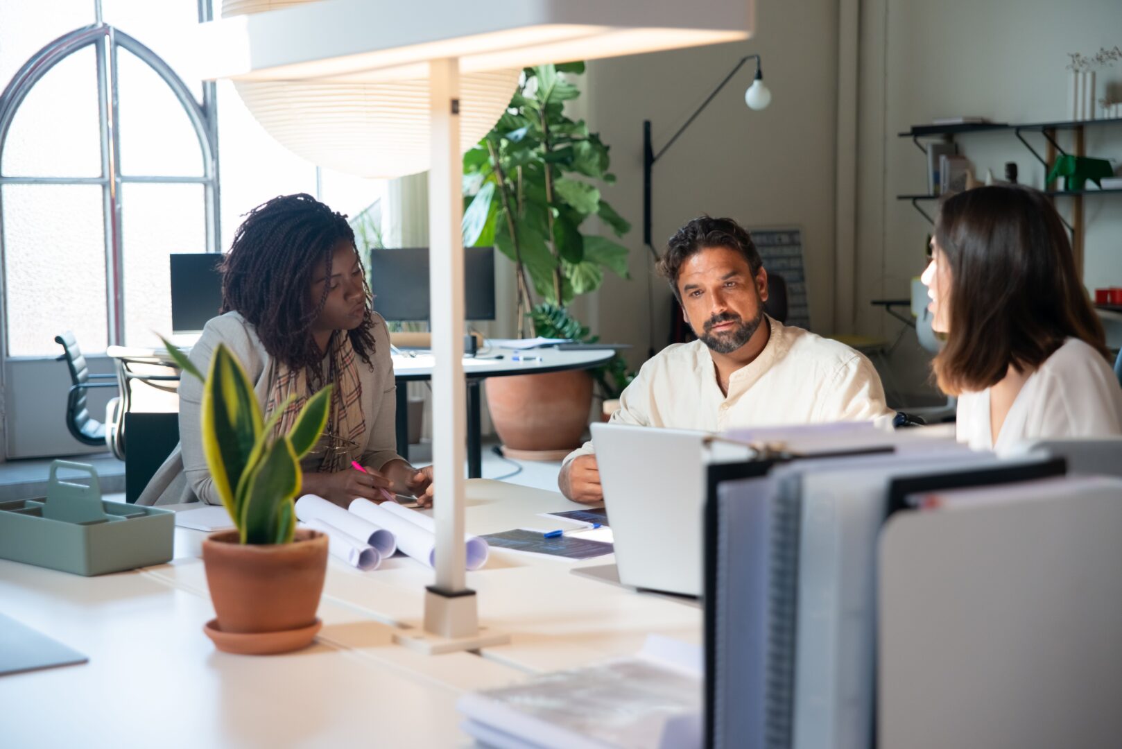 Three colleagues in discussion at an office desk, with laptops, papers, and plants in a bright workspace.