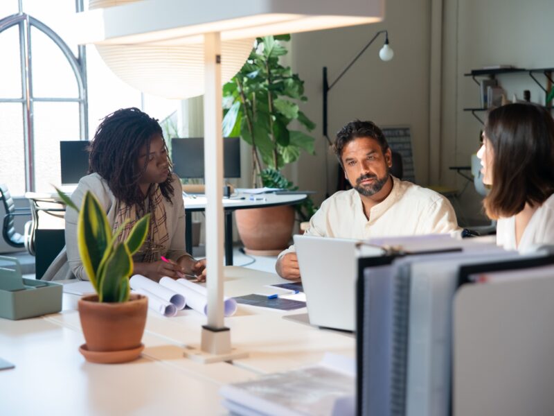 Three colleagues in discussion at an office desk, with laptops, papers, and plants in a bright workspace.