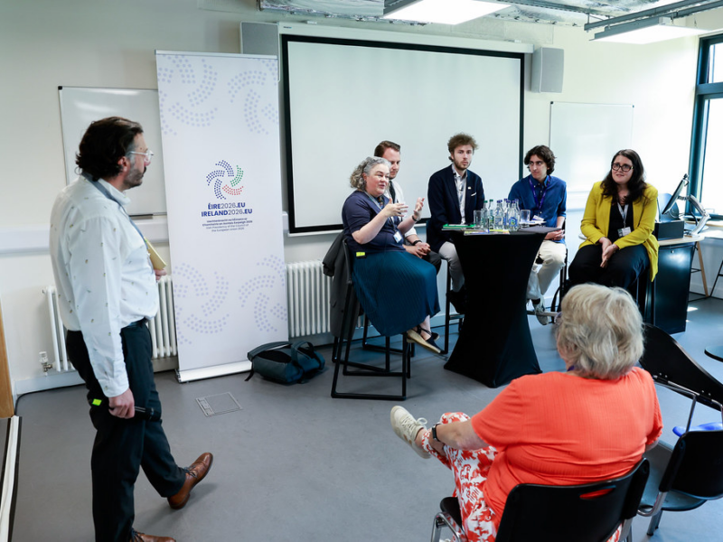A panel of five people sitting on high chairs around a round table in front of a screen and talking to the audience. A woman sitting in the audience is shown from behind while a man with a microphone in hand is standing to the side of the panel. A roll-up with the logo of the Irish Presidency of the Council of the European Union standing next to the screen.