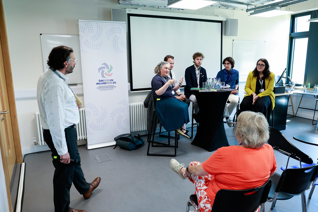 A panel of five people sitting on high chairs around a round table in front of a screen and talking to the audience. A woman sitting in the audience is shown from behind while a man with a microphone in hand is standing to the side of the panel. A roll-up with the logo of the Irish Presidency of the Council of the European Union standing next to the screen.