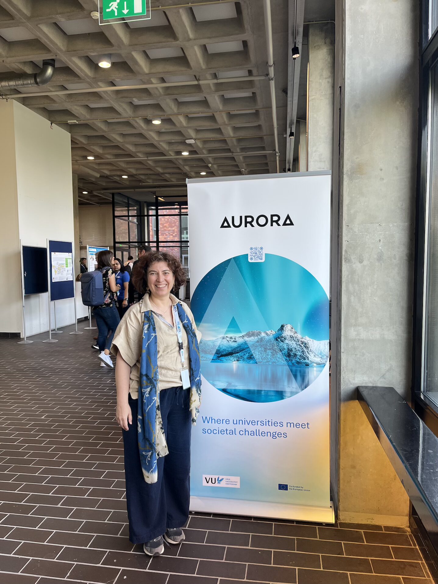Prof. İlke Şanlıer smiles while standing next to an Aurora roll-up banner reading “Where universities meet societal challenges” in VUA hallway.