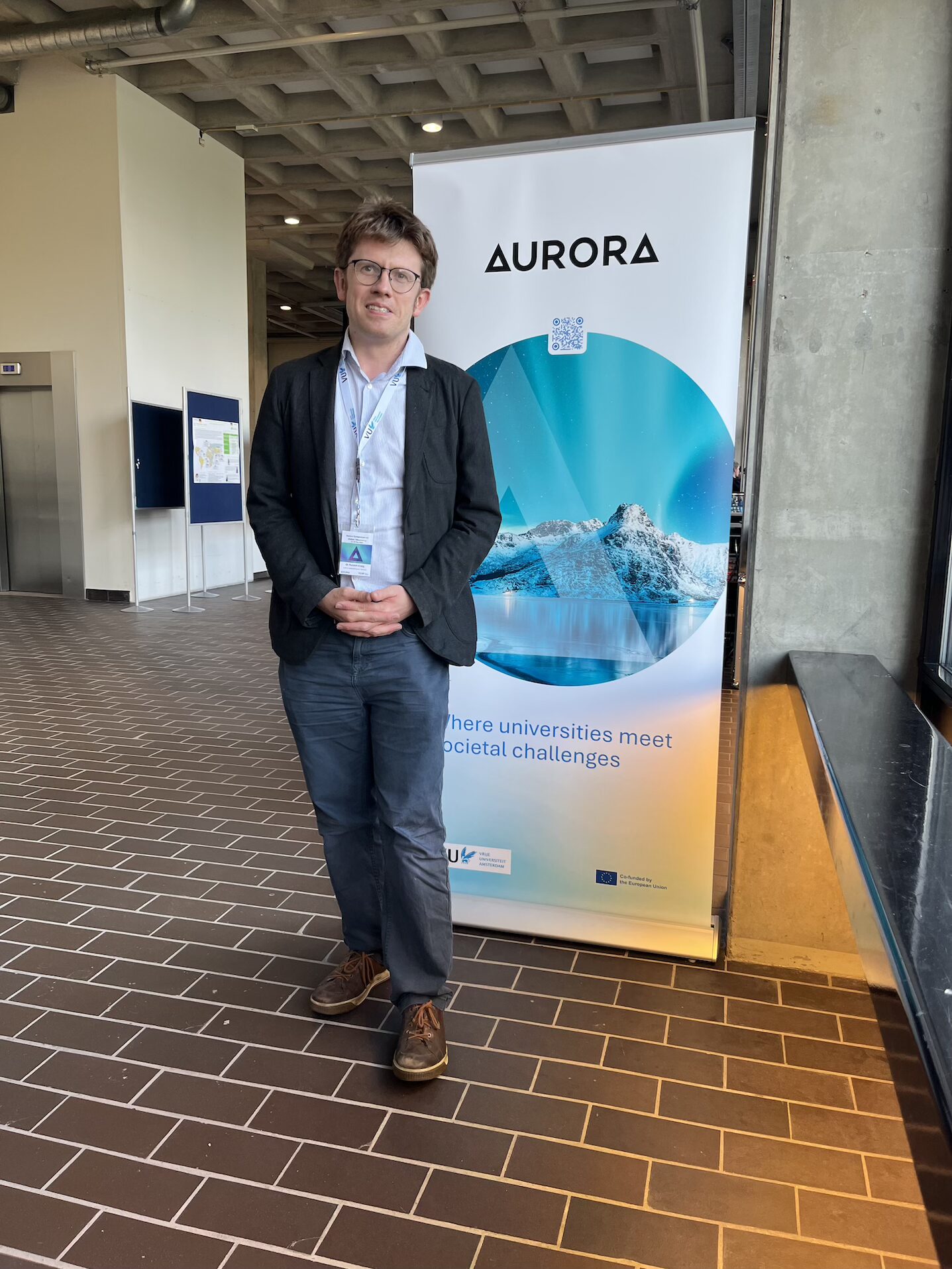 Dr. Rob Craig stands in front of an Aurora roll-up banner reading “Where universities meet societal challenges” in VUA hallway.