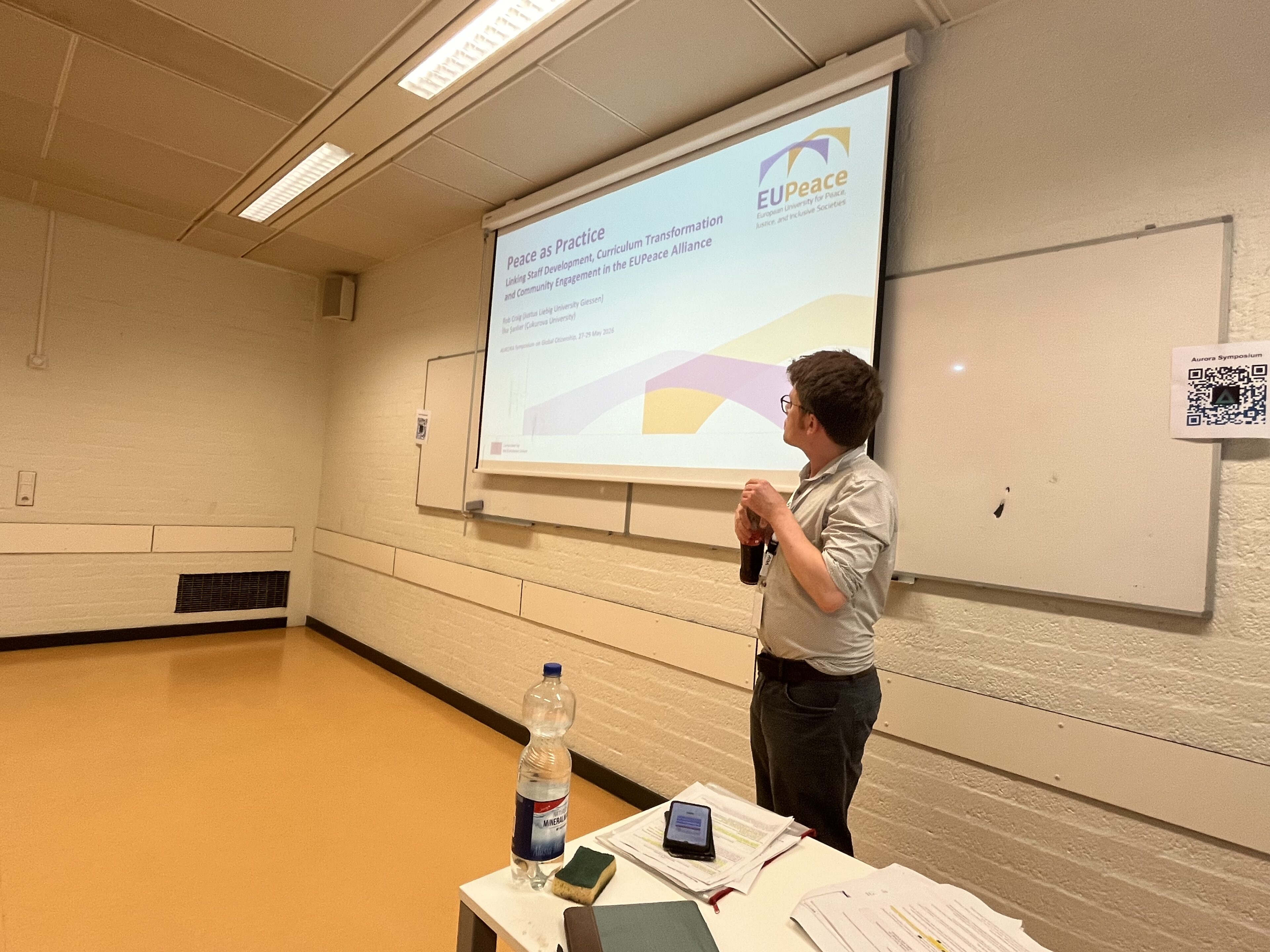 Dr. Rob Craig stands in the workshop room looking at a projected title slide reading “Peace as Practice – Linking Staff Development, Curriculum Transformation and Community Engagement in the EUPeace Alliance”