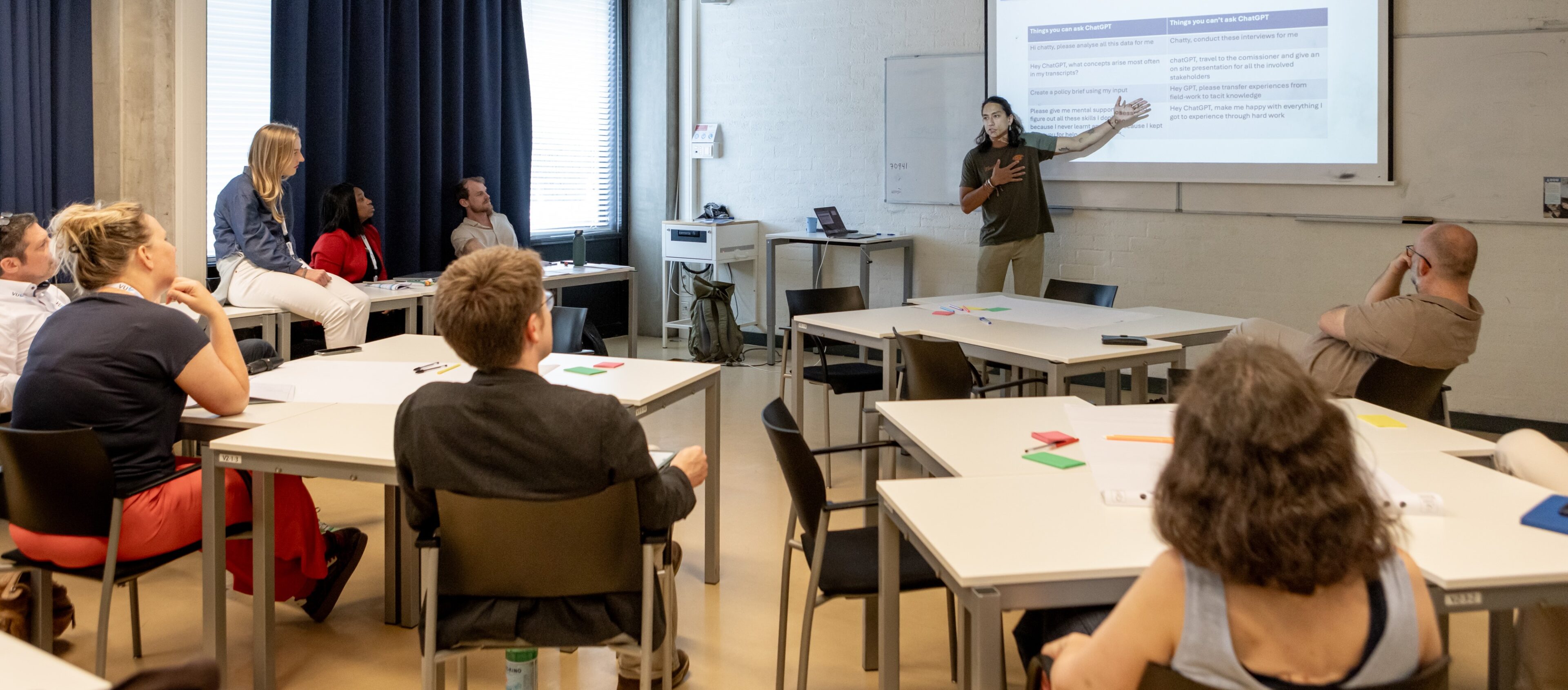 Participants seated at tables in a workshop room listen to a standing presenter gesturing at a projected slide.
