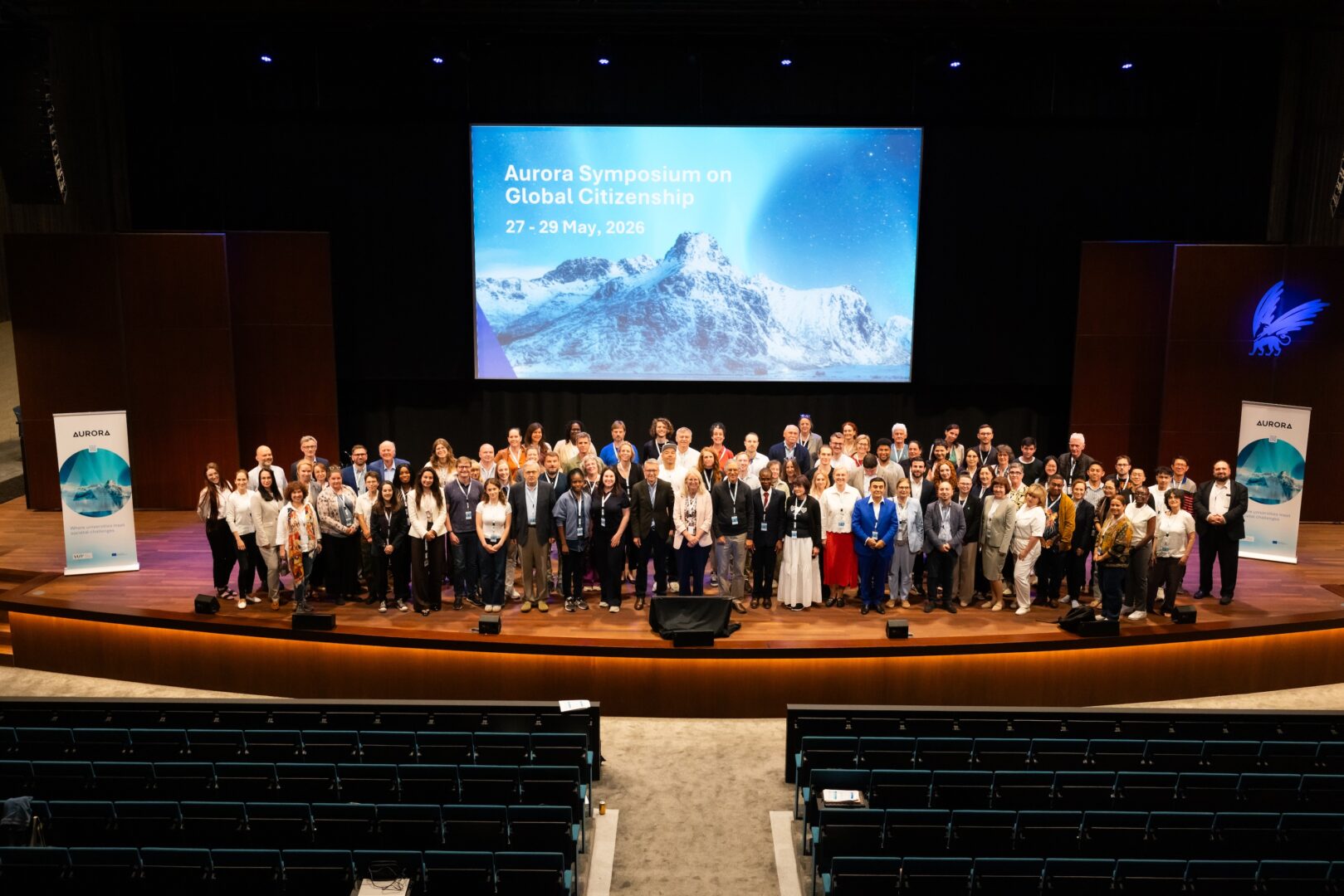 Participants of around one hundred conference participants poses on an auditorium stage beneath a screen reading “Aurora Symposium on Global Citizenship, 27–29 May 2026,” flanked by Aurora banners.