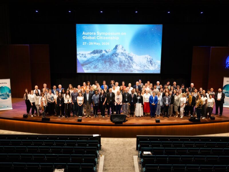 Participants of around one hundred conference participants poses on an auditorium stage beneath a screen reading “Aurora Symposium on Global Citizenship, 27–29 May 2026,” flanked by Aurora banners.