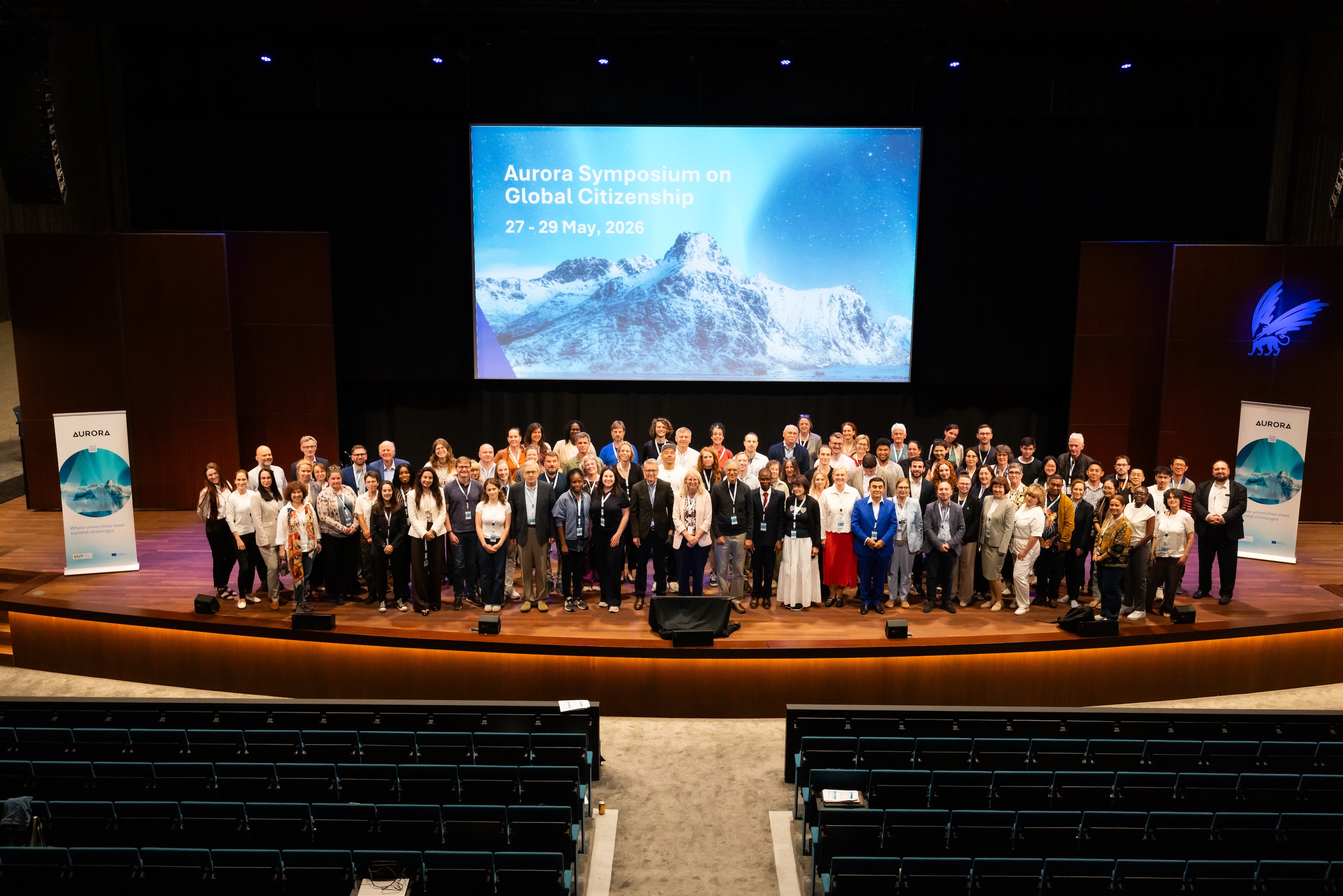 Participants of around one hundred conference participants poses on an auditorium stage beneath a screen reading “Aurora Symposium on Global Citizenship, 27–29 May 2026,” flanked by Aurora banners.