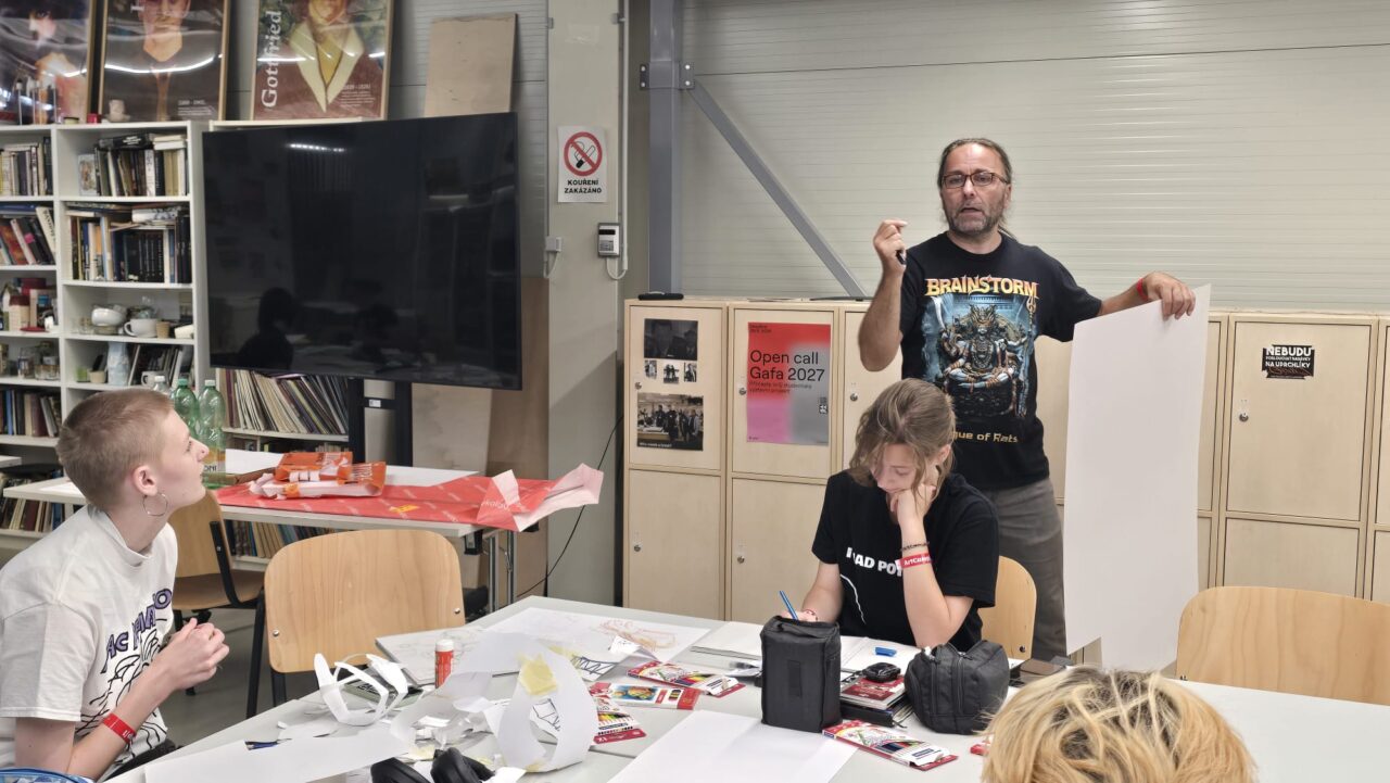 An instructor holds up a blank sheet of paper while speaking to two seated participants working on sketches at a table.