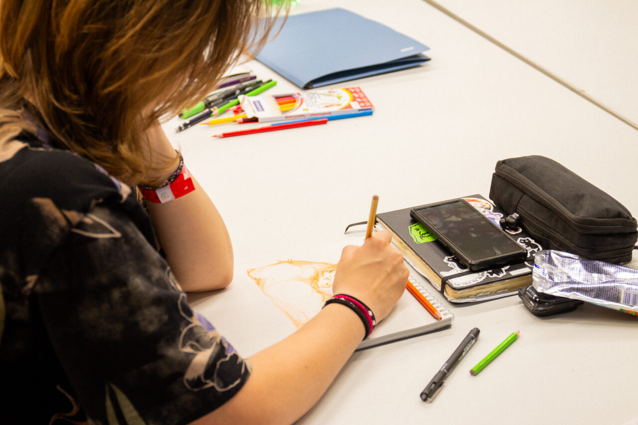 Close-up of a participant's hand sketching with a pencil in a notebook, surrounded by colored pencils and drawing supplies.