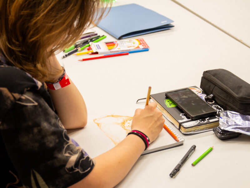 Close-up of a participant's hand sketching with a pencil in a notebook, surrounded by colored pencils and drawing supplies.