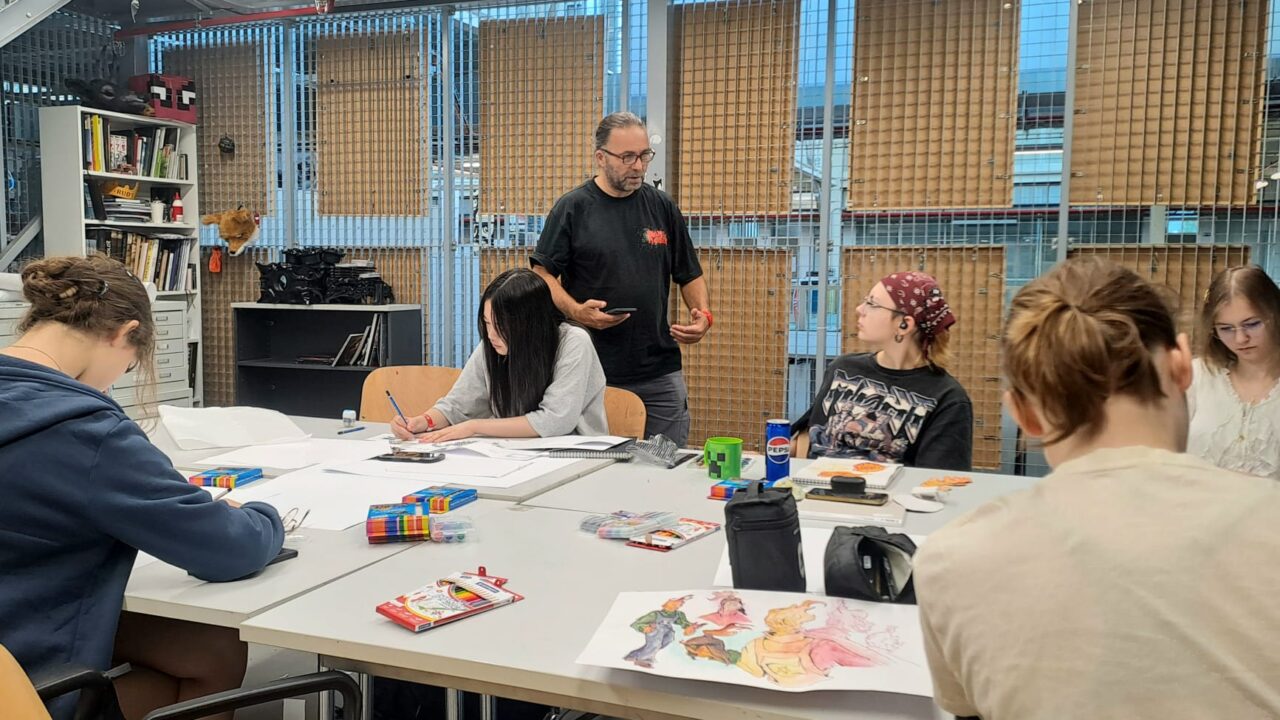 An instructor stands at a worktable talking to a group of young artists, who are drawing and sketching with colored pencils and markers spread across the table.