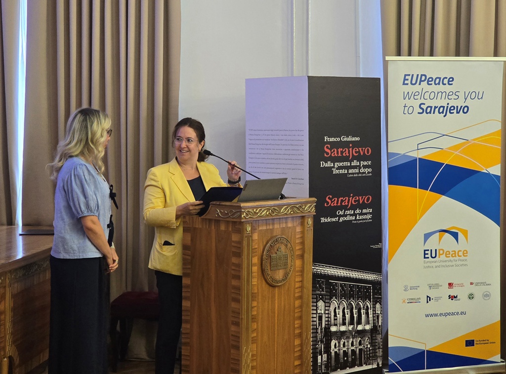 A woman standing behind a podium holding a microphone and talking to a woman standing next to her. Pillars of the EUPeace Alliance and the exhibition are located next to the podium. The woman behind the podium is Sarah Eti Castellani.