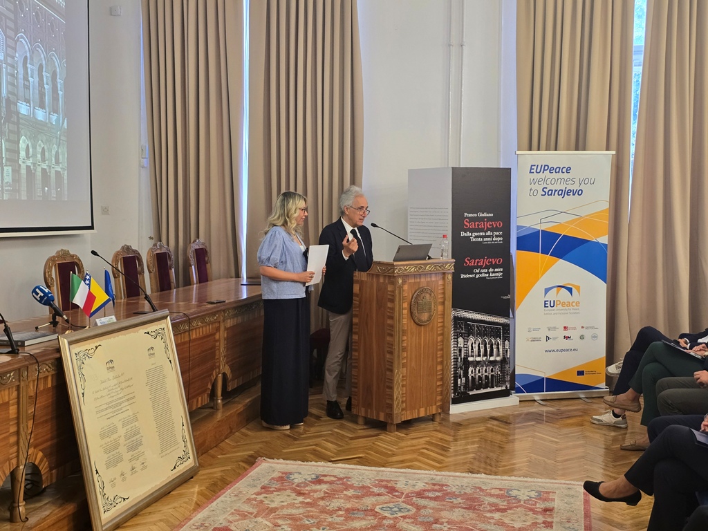 A person standing behind a podium, speaking into a microphone. The person is Franco Giualiano. A woman is standing next to him.