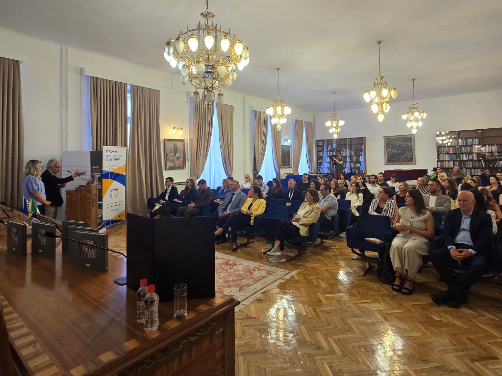The audience from the exhibition listening to Franco Giuliano, speaking behind a podium. A woman is standing next to him.