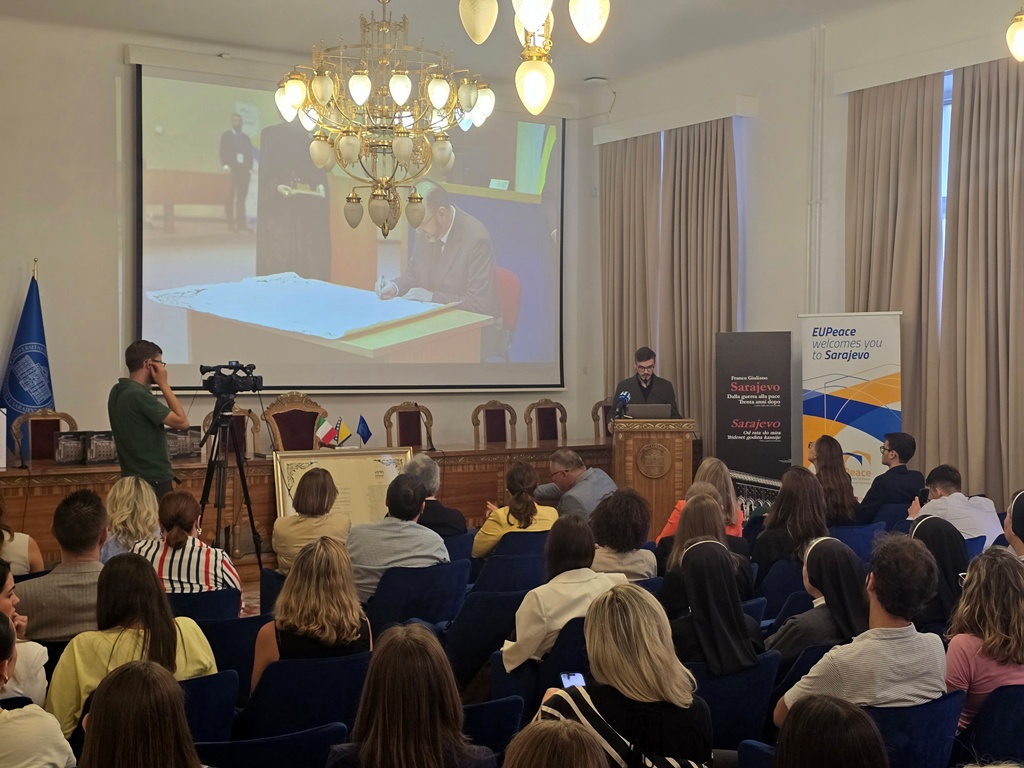 A person standing behind a podium in front of an audience. An image of a person signing a document is projected onto a screen on the wall. The person behind the podium is Imran Pašalić.