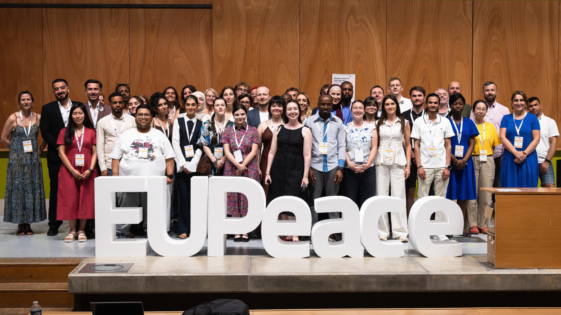 Around fifty participants of the EUPeace Doctoral Training Week stand on an auditorium stage behind large white three-dimensional letters spelling "EUPeace".