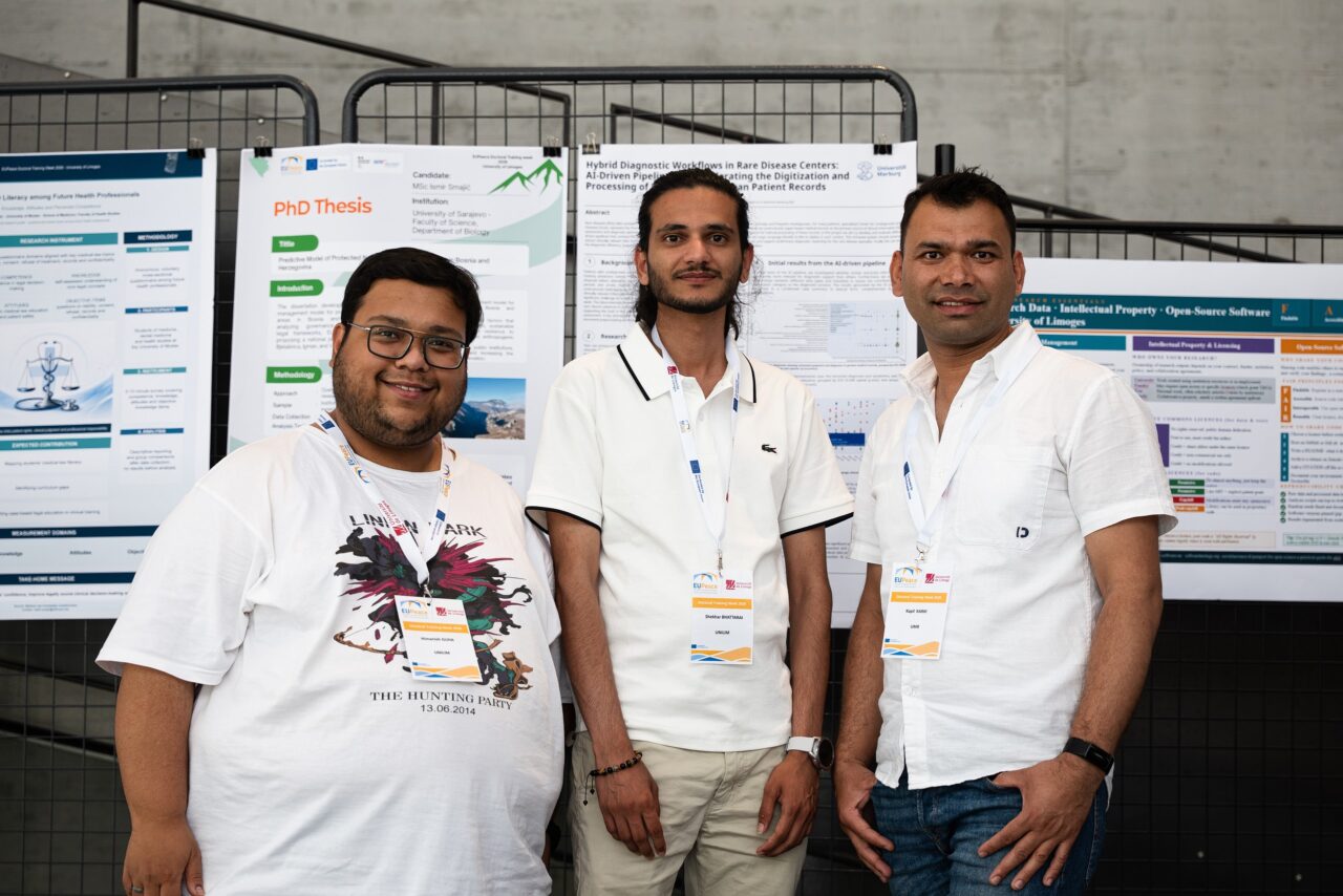 Three doctoral candidates smile at the camera in front of a row of research posters, wearing EUPeace Doctoral Training Week lanyards.