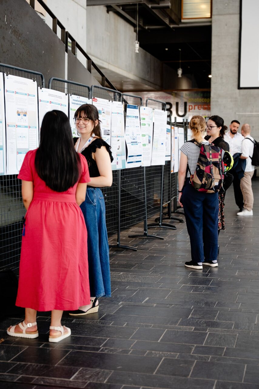 Participants stand in small groups in a university foyer, discussing research posters displayed on metal panels.