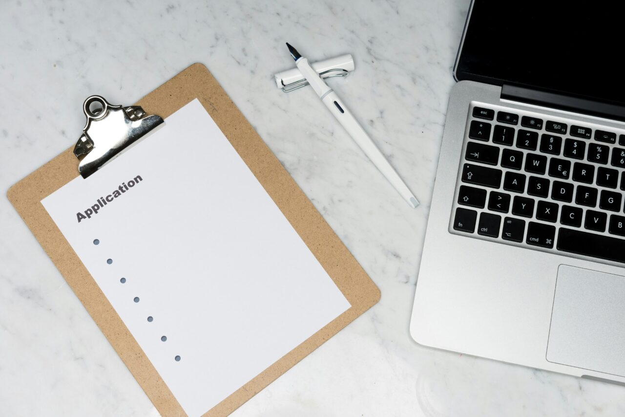 A clipboard with a blank application form on a marble desk, next to a pen and an open laptop.