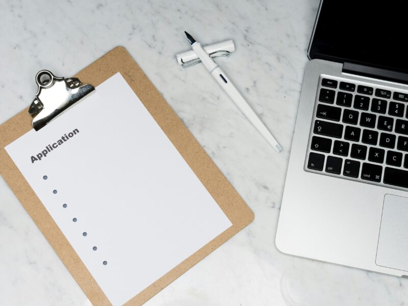 A clipboard with a blank application form on a marble desk, next to a pen and an open laptop.
