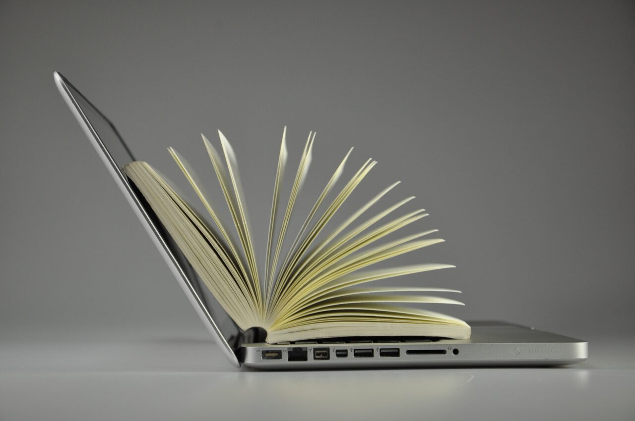 A photo illustration of an open book with fanned pages merging seamlessly into an open laptop, side view against a grey background.
