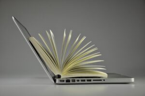 A photo illustration of an open book with fanned pages merging seamlessly into an open laptop, side view against a grey background.