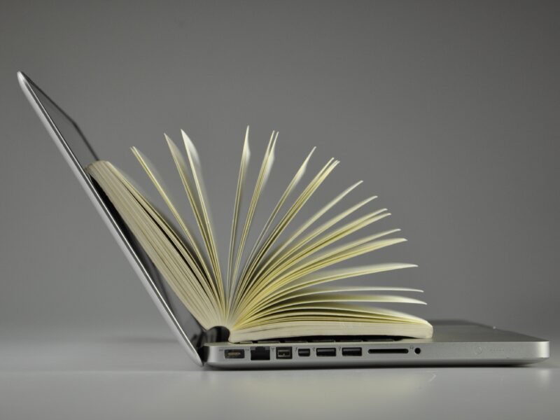 A photo illustration of an open book with fanned pages merging seamlessly into an open laptop, side view against a grey background.