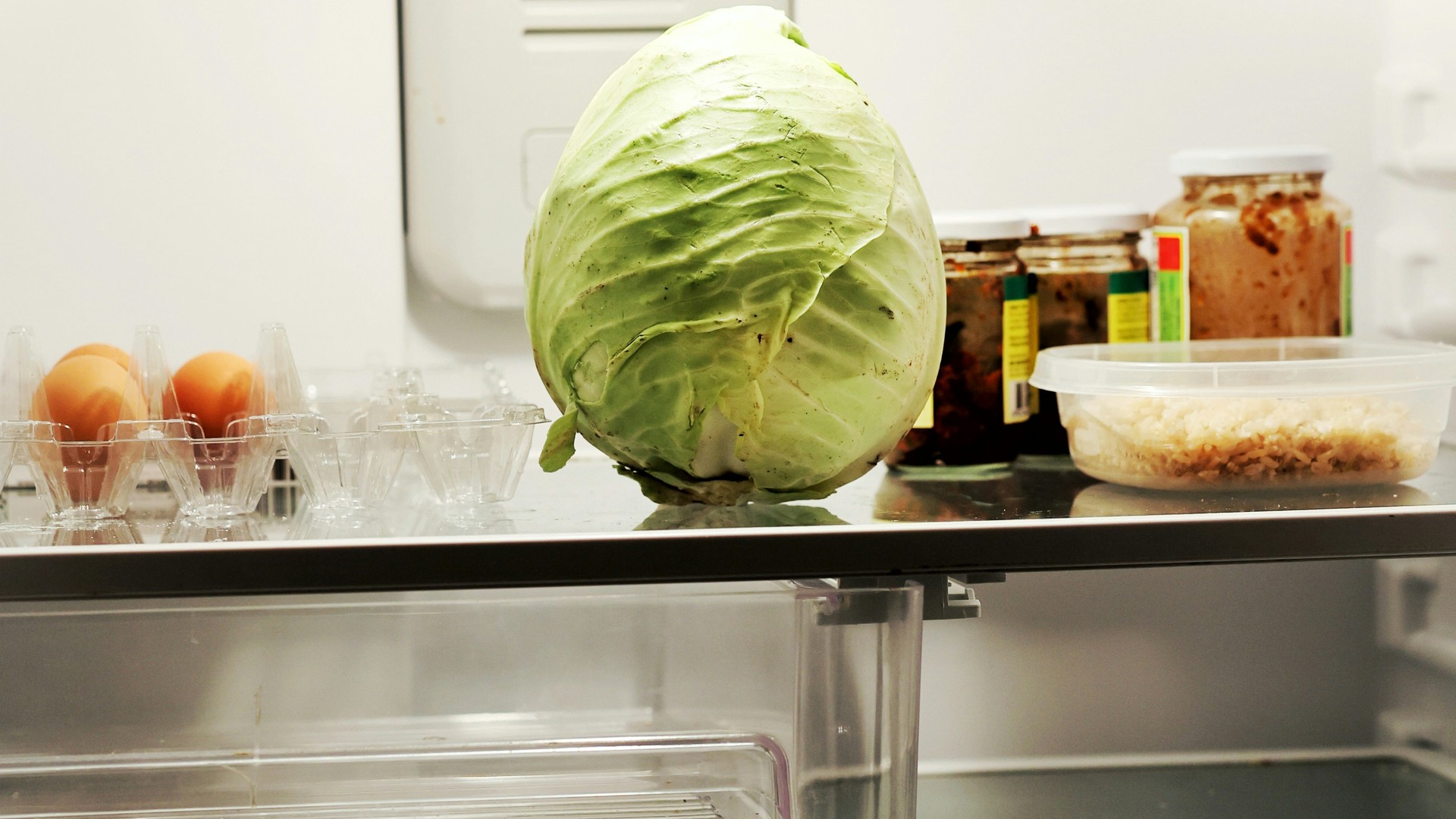 View into an open, sparsely stocked fridge. On a glass shelf sit a single head of green cabbage, an egg tray holding only two eggs, a few jars of preserved food and a plastic container of rice. The drawer beneath the shelf is empty.